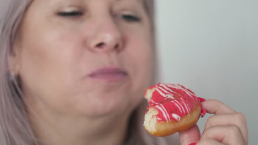 Close-up of a middle-aged Caucasian woman enjoying a sweet round donut with icing, woman eating dessert, slow motion