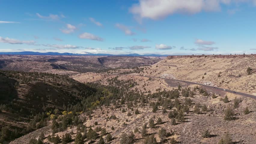 Aerial view of U.S. Highway 26 winding through a landscape of canyons and sparse vegetation under a blue sky with scattered clouds, Warm Springs, Oregon, United States.