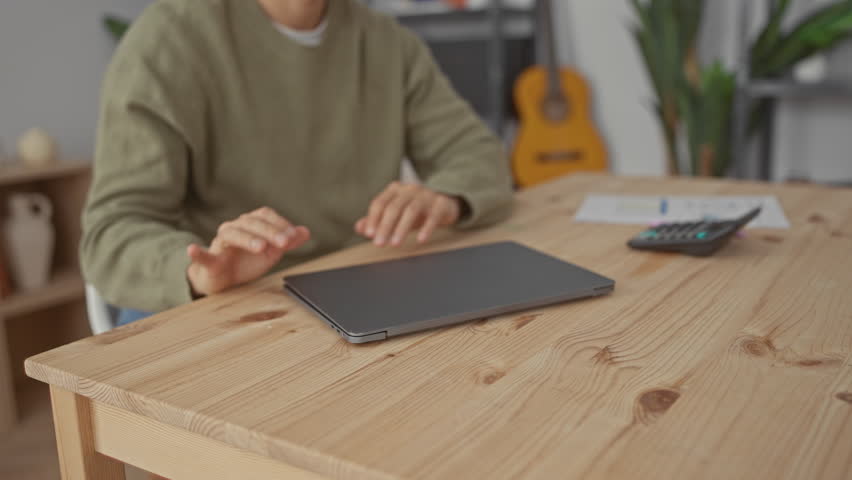 Young man working from home on a laptop in a cozy living room featuring plants and a guitar, focusing on a financial spreadsheet with a calculator nearby on the wooden table.