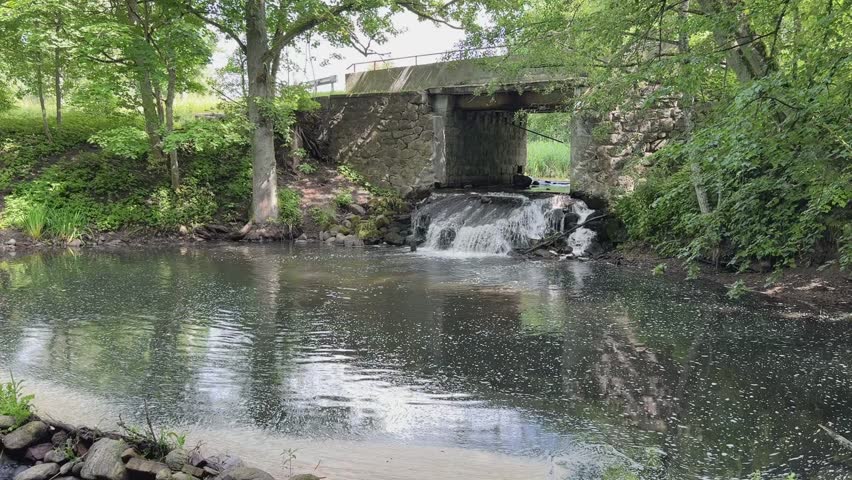 Strong water flow under old dam or small hydro structure, water splashing over rocks, surrounded by green trees.
