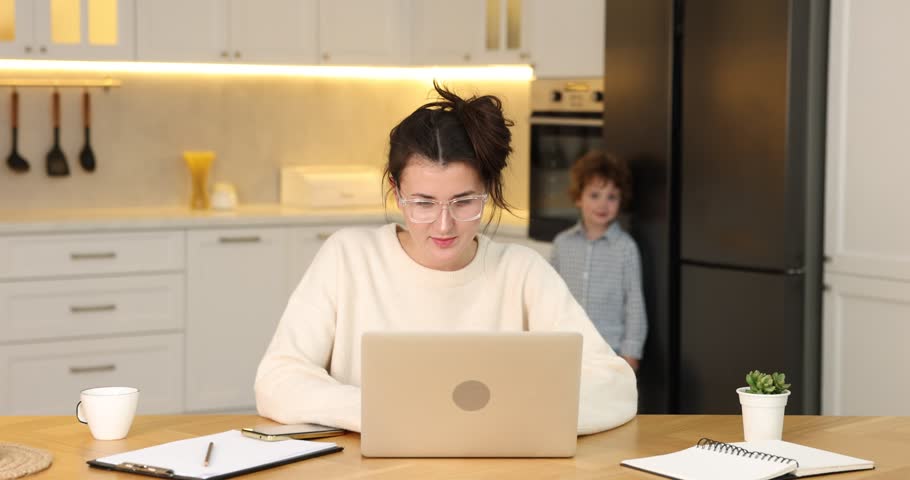Smiling woman in glasses working with laptop while her naughty son bothering her at wooden desk in home office