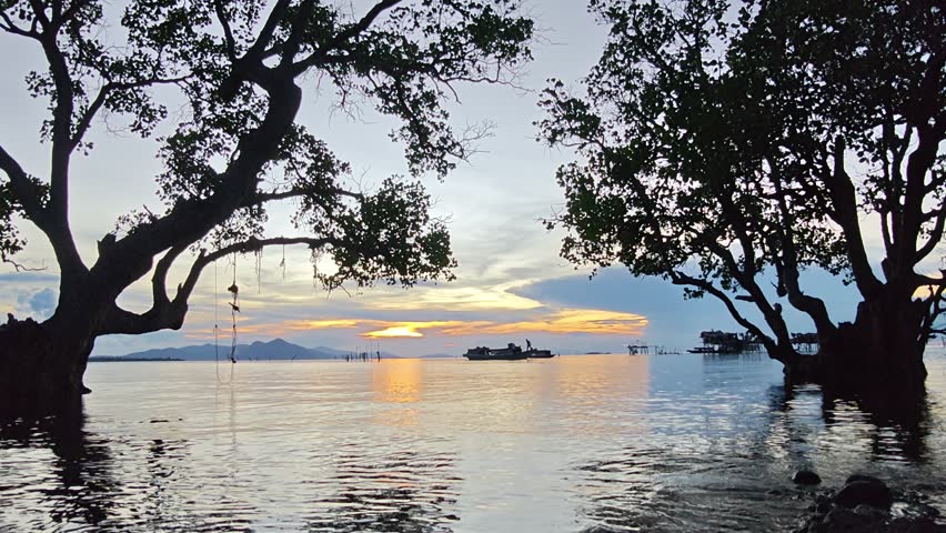 Beautiful sunset over the calm sea with silhouette trees and fishing boats in Semporna, Sabah, Malaysia. Tranquil tropical seascape captured in a peaceful evening timelapse.
