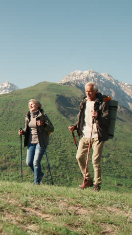 Slow motion vertical video elderly couple hiking with poles in mountains active lifestyle wellness healthy aging fitness retirement outdoor activity freedom longevity wellbeing nature