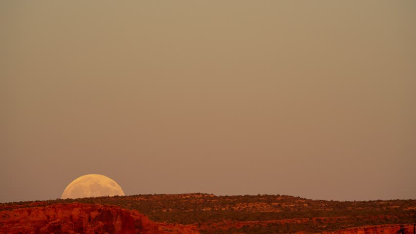 Timelapse of a large full moon rising over desert terrain as the sun is setting