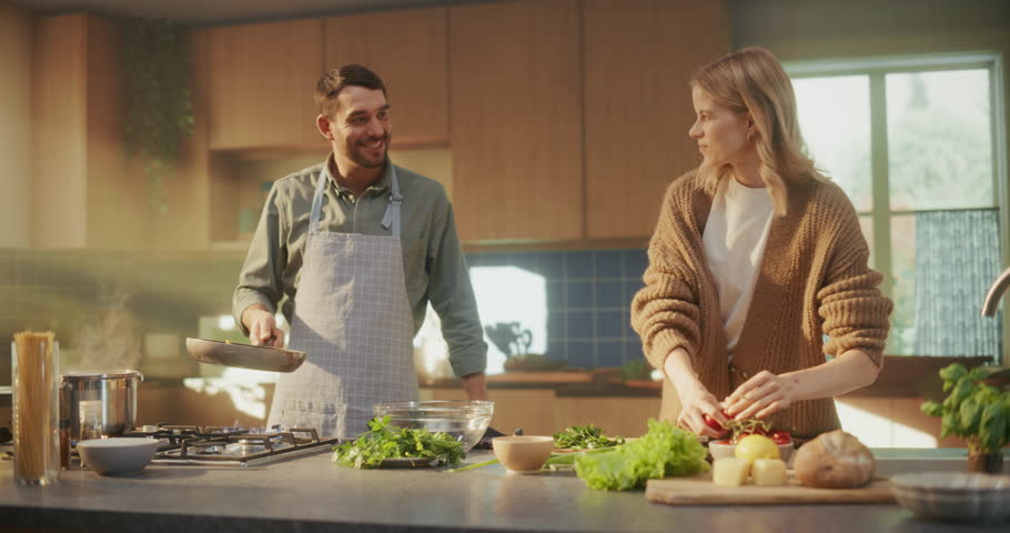 Modern Family Couple Spending Quality Time in the Kitchen. Man in Apron Prepares Dinner on the Stove, Female Helps with the Cooking, Standing By His Side, Cutting Fresh Vegetables