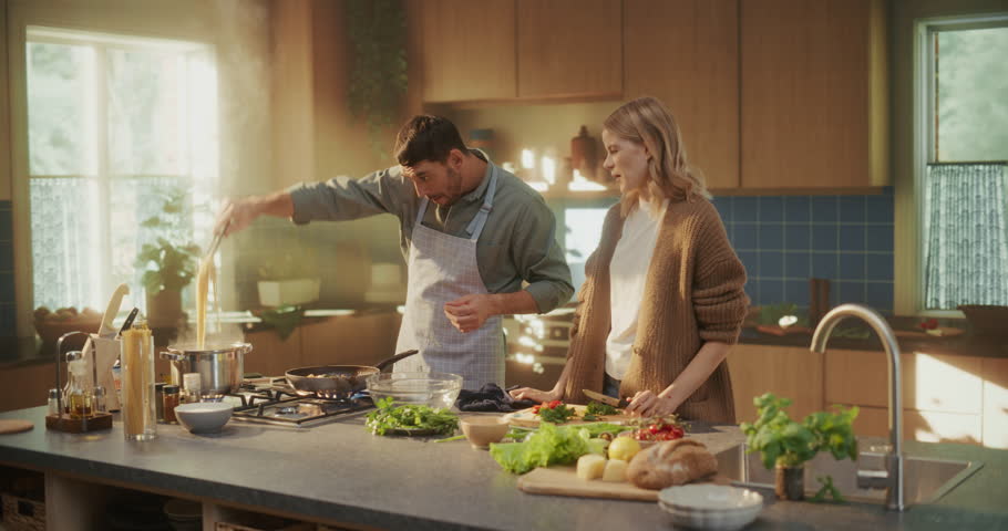 Young Couple Cooking Together in a Bright Modern Kitchen. Man in Apron Boils Pasta and Cooks Shrimps while His Girlfriend Prepares a Fresh Salad. Family Sharing a Relaxed and Joyful Day at Home