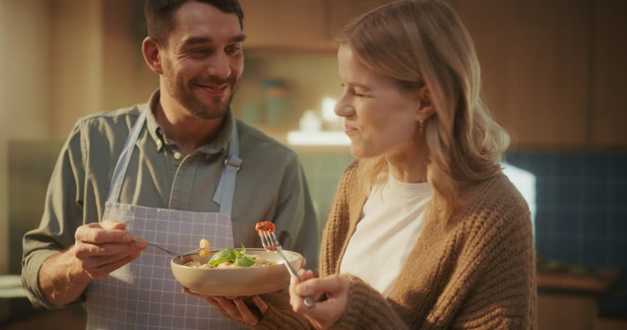 Caucasian Couple Tastes Pasta in a Cozy Kitchen. Man, Wearing an Apron, Shares a Bowl with His Girlfriend or Wife. Young Family Couple Enjoy the Delicious Italian Meal