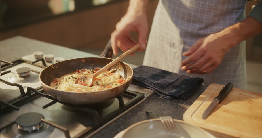 Caucasian Man Stirring Pasta in a Frying Pan and Serving It on Plate in a Cozy Kitchen. His Beautiful Wife Standing Beside Him and Adding a Basil Leaf to Finish the Dish