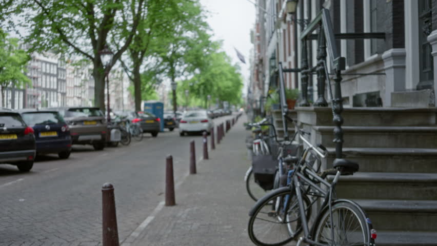 Defocused view of an amsterdam street with bikes leaning against a building and parked cars lining the tree-shaded road.