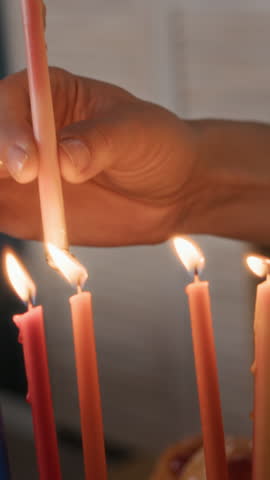 Vertical cropped slowmo of hands of unrecognizable woman putting burning shammash on hanukkiah, preparing festive table for Hanukkah celebration at home