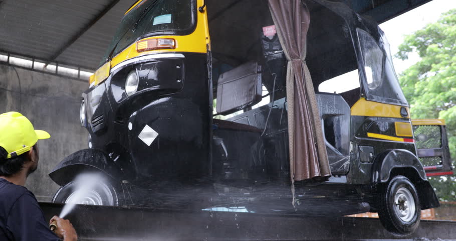A black and yellow auto rickshaw being washed using a water pipe at a local service area in Navi Mumbai, India. The video shows daily vehicle cleaning work with splashing water 