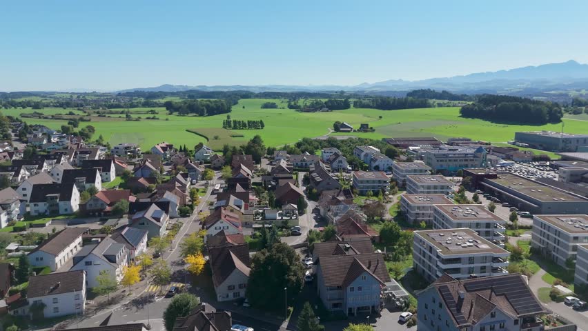 Modern Swiss town neighborhood with solar panels on houses. Rural farm fields in background at sunny summer day. City of Zuzwil in Switzerland with alps in distance. Aerial dolly wide.
