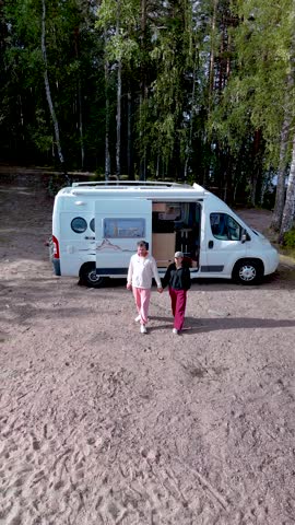 A couple of men and women stroll along the sandy shore of Lake Saimaa Finland, surrounded by lush greenery and a tranquil atmosphere. A camper van is parked on the beach