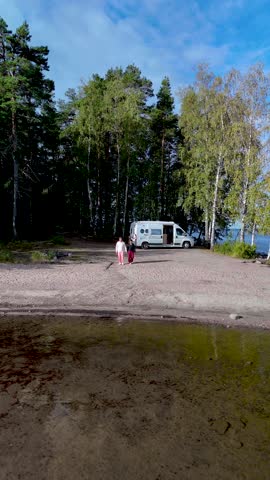A couple of men and women stroll along the sandy shore of Lake Saimaa Finland, surrounded by lush greenery and a tranquil atmosphere. A camper van is parked on the beach
