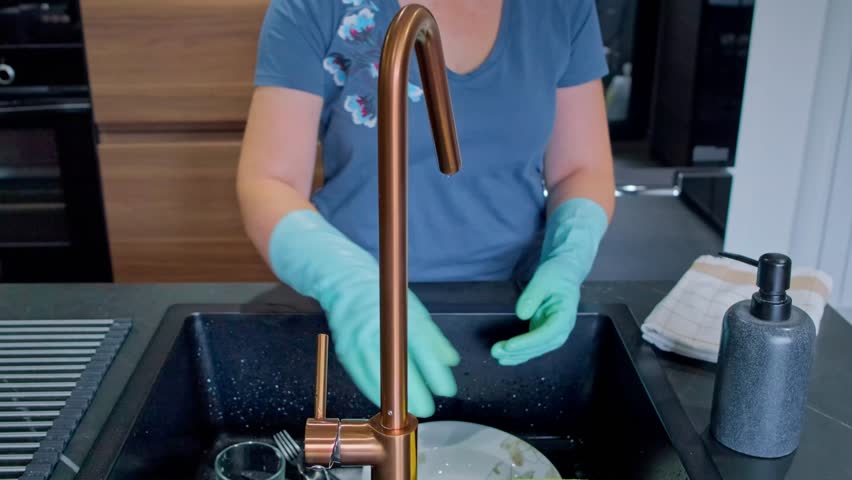 Unrecognizable woman wearing protective rubber gloves using a sponge and soap to clean dirty dishes, glasses and cutlery in a modern kitchen black sink, performing a common household chore