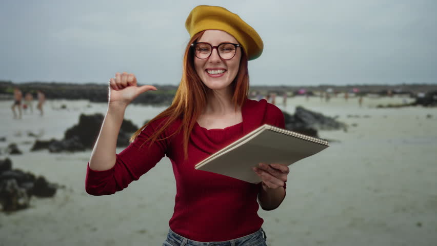 Woman with red hair smiling and pointing at herself on a sandy beach with a sketchbook, wearing a yellow beret and glasses outdoors at the seaside.