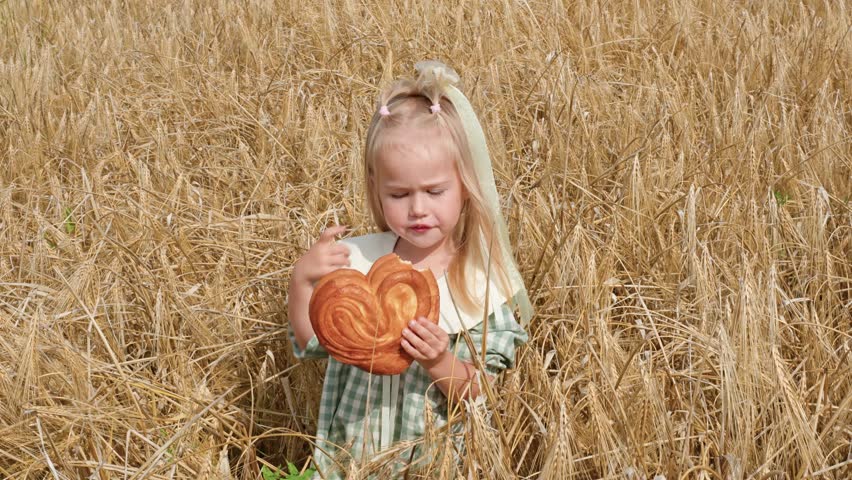 A little child girl in dress in a field of wheat eats a bun in summer, the concept of a happy healthy childhood in the countryside