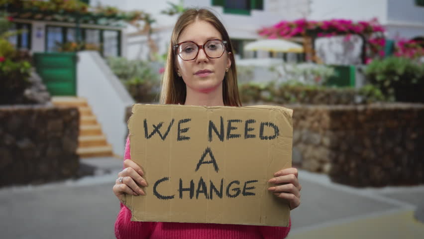 Young woman with blonde hair and glasses holding cardboard sign with bare hands on street; progress determination.