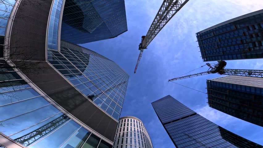 Dynamic low angle view of modern glass skyscrapers and construction cranes against a bright blue sky, showcasing urban development, contemporary architecture, and corporate real estate growth