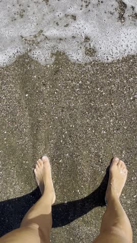 POV shot of bare female feet in foamy waves on dark sand beach as camera slowly moves forward into the sea toward the horizon