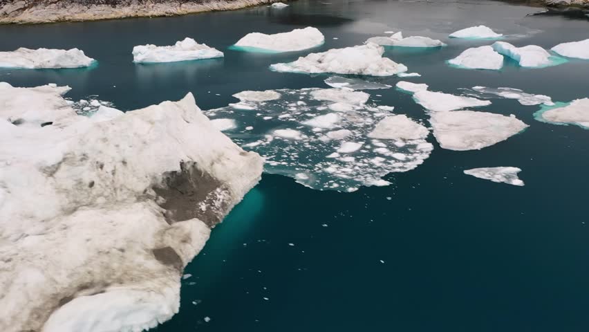 Aerial drone view of floating icebergs drifting in a calm Arctic bay surrounded by rugged rocky hills in Greenland, showcasing the contrast between icy water and barren landscape.