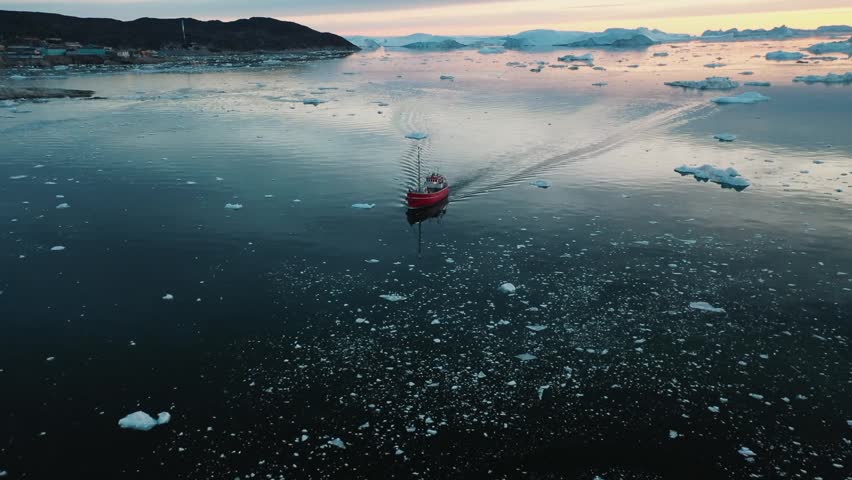 Aerial view of a small red fishing boat moving through icy Arctic waters in Greenland at sunset, with floating icebergs and colorful reflections creating a peaceful polar scene.