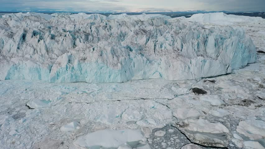 Aerial drone view of a towering glacier wall surrounded by a frozen landscape in Greenland, showcasing the dramatic beauty and scale of the Arctic environment.