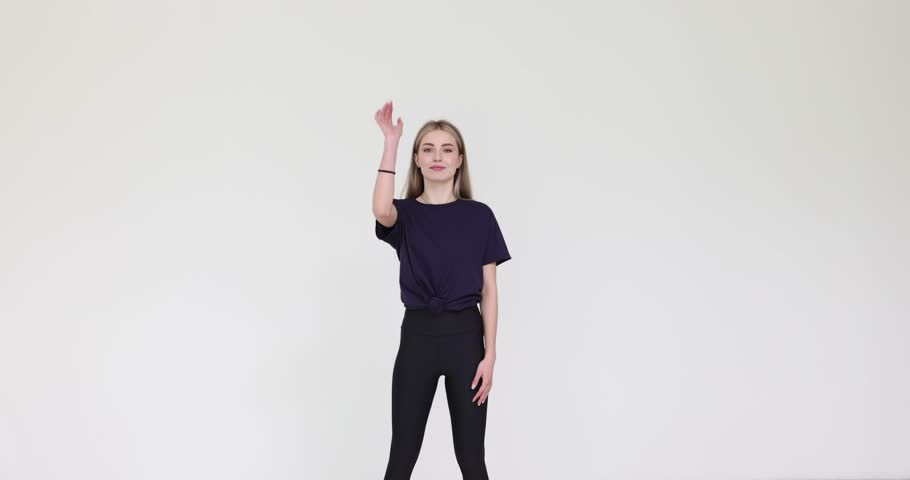 Young woman shows simple dance moves by arms on white background. Blonde female in black activewear rehearses performance indoors slow motion
