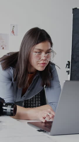 Vertical shot of professional photographer leaning on table and looking at screen while working on laptop and editing photos
