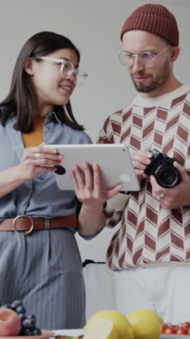 Vertical shot of two Caucasian photographers working together in studio. Brunette woman holding tablet, showing reference photos while man holding camera and listening to her speech