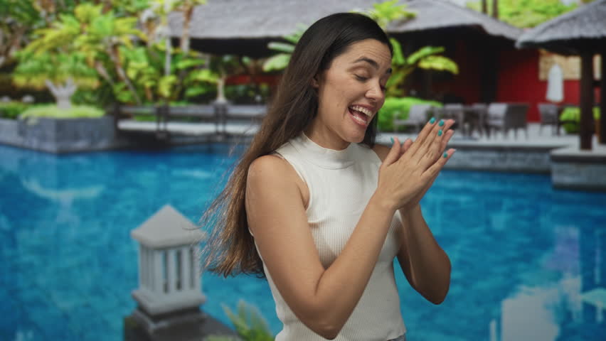 Woman rubbing hands beside resort pool building, sleeveless top and teal nails, smiling with eyes closed and leaning forward; excitement vacation.