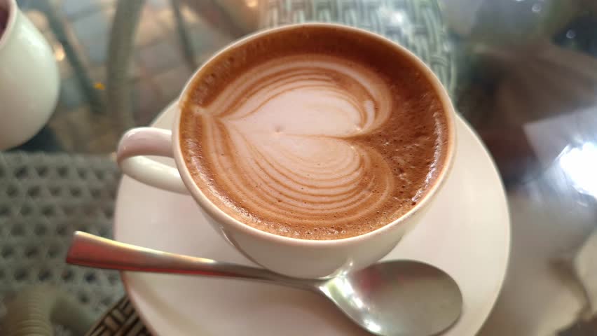 Warm cup of cappuccino with heart-shaped latte art and spoon placed on a saucer, perfect for café, breakfast, or relaxation themes.
