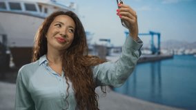 Woman holding smartphone at port taking a selfie, hand on cheek and smiling with windblown hair; carefree joy. - Powered by Shutterstock - Get 15% off with code: PIKWIZARD15