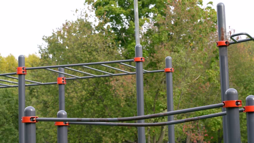 Modern outdoor calisthenics structure with gray metal bars and orange accents, set in lush green park, ready for strength training and bodyweight workouts.