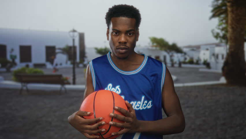 Man wearing blue basketball jersey holds basketball on sunny street beside wooden bench; confidence aspiration.