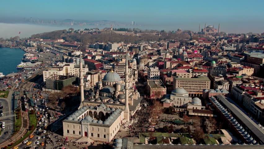 Aerial panorama of Istanbul’s historic peninsula where the Yeni Mosque dominates Eminonu, with morning fog over the Bosphorus revealing Topkapi Palace and Hagia Sophia in the distance.