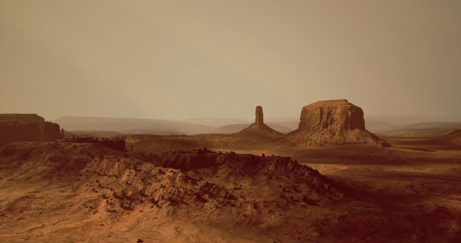 A sweeping view of a barren desert features towering rock formations and rolling hills. The atmosphere is hazy, hinting at a stunning sunset illuminating the landscape.