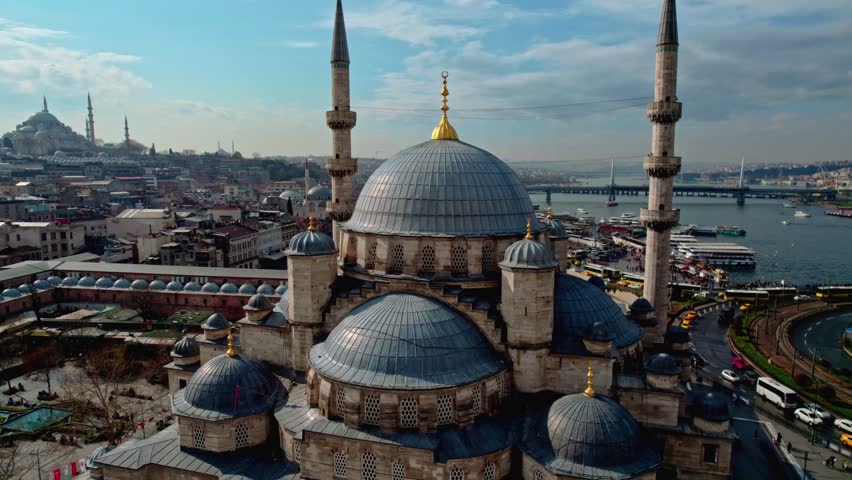 Drone footage of Istanbul showing the New Mosque, Eminonu Square, Suleymaniye Mosque, and the Golden Horn. Historic cityscape with Bosphorus and Ottoman architecture under blue sky.