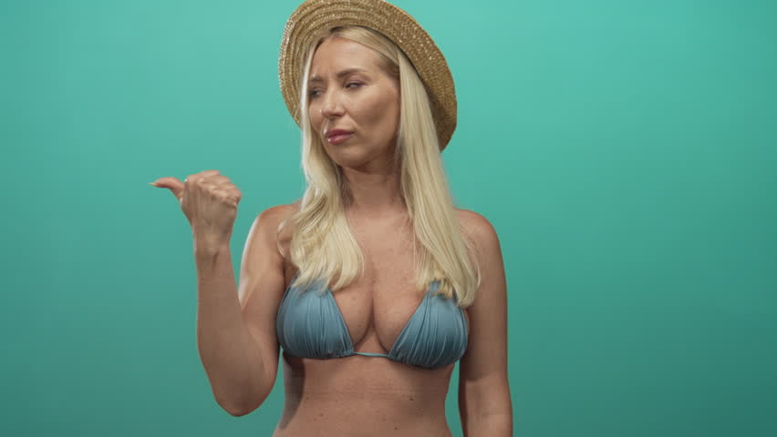 Blonde woman in a turquoise studio wearing straw hat and bikini top points thumb backward at air; skepticism.