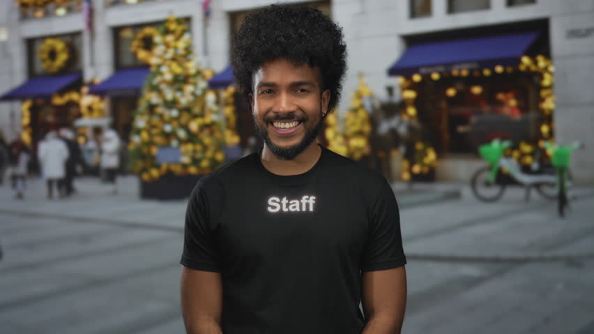 Man with afro hairstyle in staff uniform smiling outside on a decorated city street with festive lights and bicycles visible in the background.