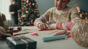 Focused African American girl child drawing on Christmas greeting card or writing letter to Santa Claus with colorful pencils while crafting DIYs with family in living room - Powered by Shutterstock - Get 15% off with code: PIKWIZARD15