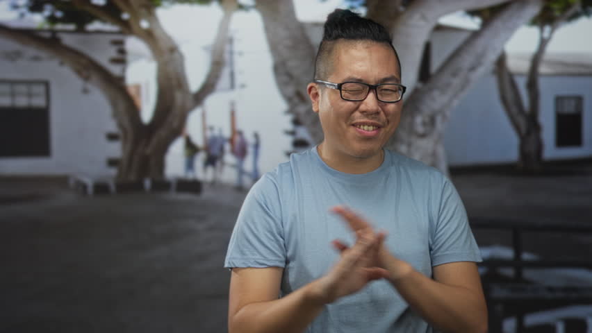 Young chinese man smiling with hands raised, wearing glasses and a blue tshirt while seated on a bench on a street in front of a white building under large trees; joy warmth.