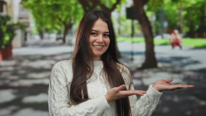 Woman smiling with outstretched arms in outdoor urban setting surrounded by lush greenery, captured on a sunny street in a lively city environment.
