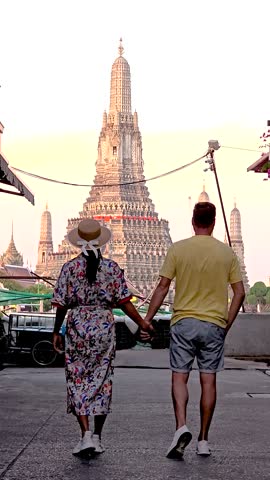 A couple enjoys a romantic walk hand in hand while approaching the iconic Wat Arun temple at sunset. The golden hues create a magical atmosphere in the vibrant city of Bangkok.