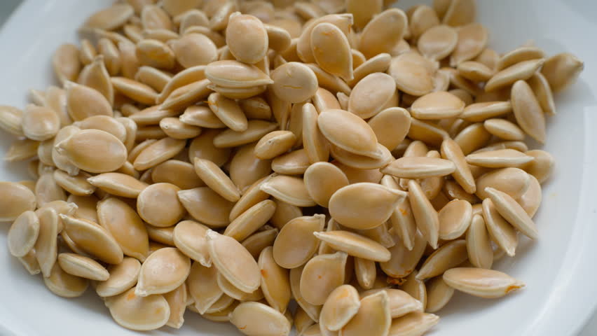 Close up shot of a hand picking up one raw shelled pumpkin seed from a white bowl full of seeds