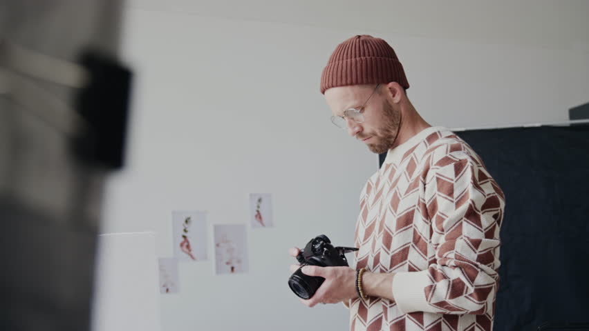 Adult bearded man holding professional camera in hands and looking at new food shots, he standing in modern studio with photos on walls and photography equipment