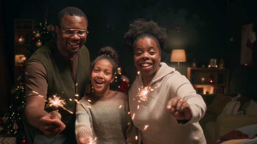 Portrait of smiling Black girl child holding sparkling Bengal flares next to mom and dad while having fun together in living room on Christmas Eve, slow motion