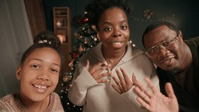 Cheerful African American family of two adults and girl child waving hands saying hello in video call with relatives while showing cozy living room decorated with Christmas tree - Powered by Shutterstock - Get 15% off with code: PIKWIZARD15