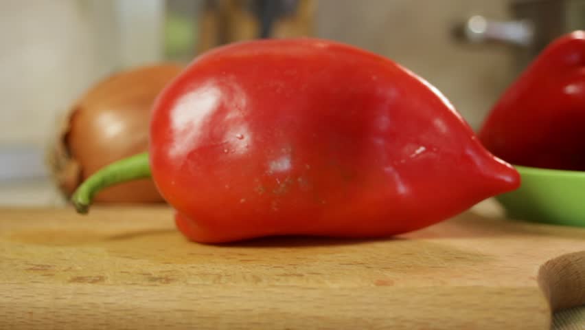 Male hands cut ripe red bell pepper with kitchen knife on wooden cutting board surrounded by fresh vegetables. Slice reveals natural texture. Concept of homemade cooking and vegetable preparation