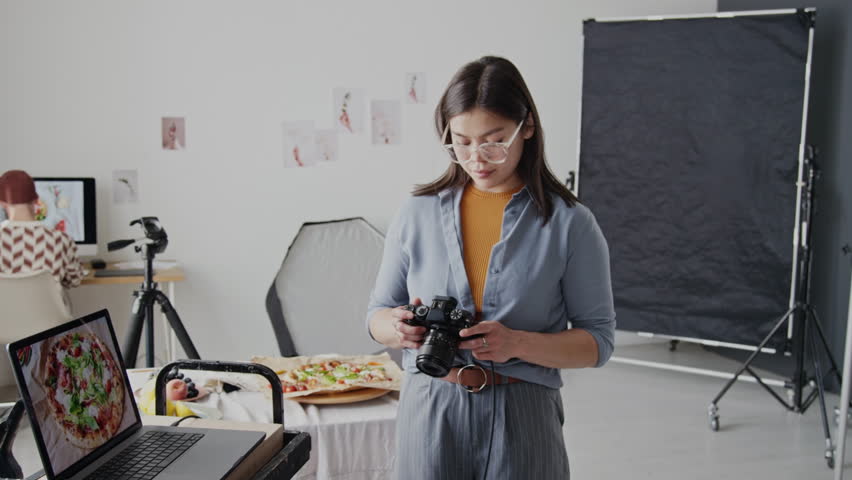Young female photographer holding professional camera while posing and smiling, adjusting glasses